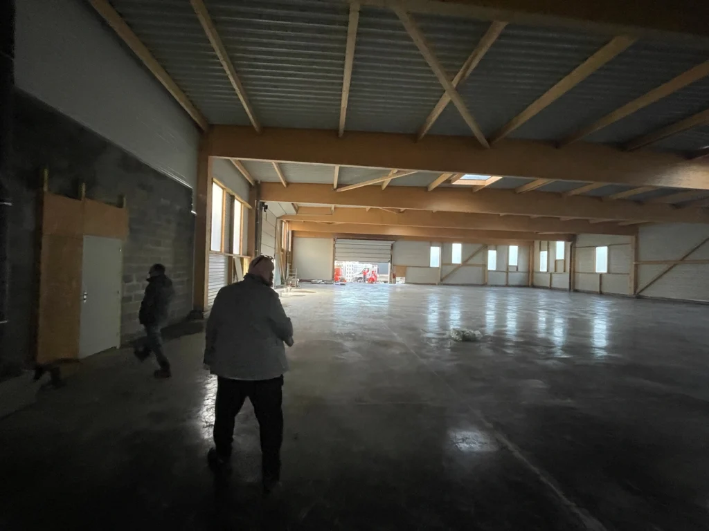 Deux personnes marchent à l'intérieur d'un grand entrepôt vide à Hadecoville, explorant un futur chantier pour des bureaux professionnels. L'espace présente des sols en béton, de hauts plafonds et des poutres apparentes, ce qui laisse présager une transformation potentielle dans le style scandinave.