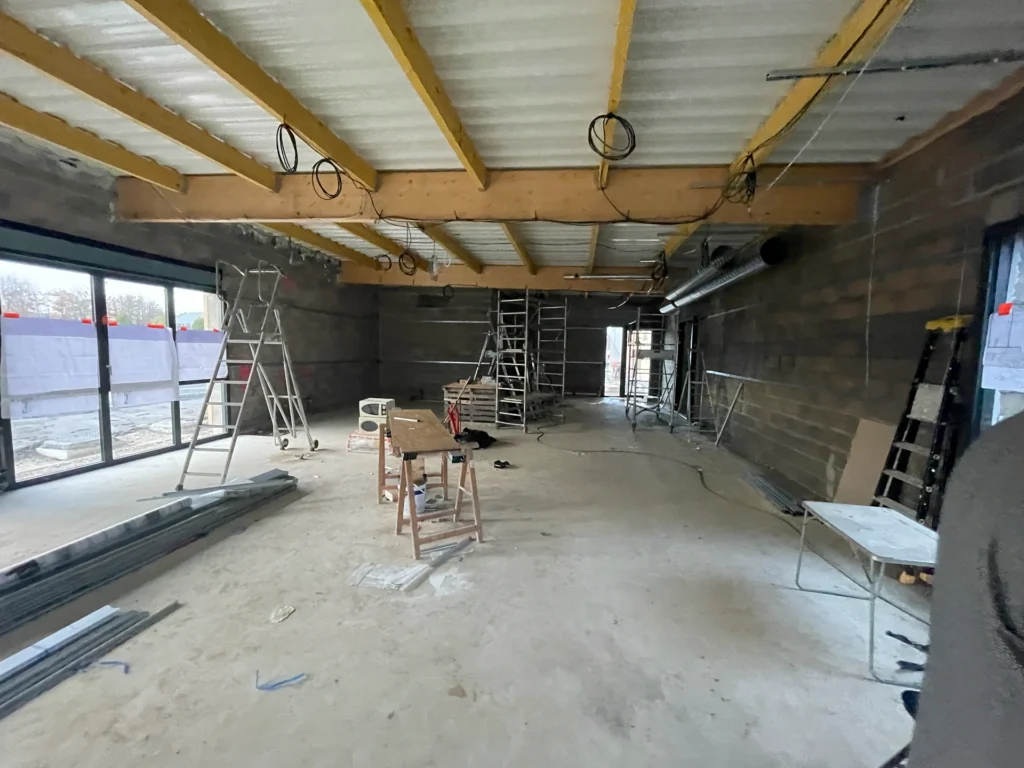 Une grande pièce en construction au Chantier Hadecoville avec des poutres apparentes, des murs inachevés, des échelles, des outils et des débris. La lumière naturelle pénètre par de grandes fenêtres, laissant entrevoir de futurs bureaux professionnels de style scandinave.