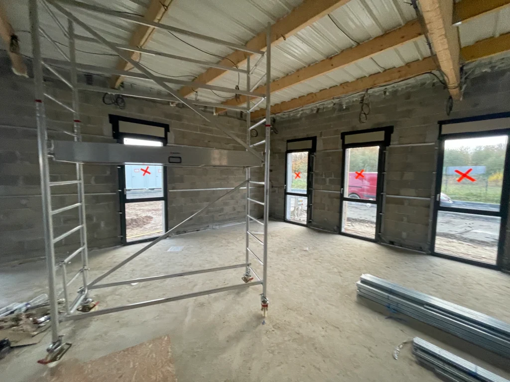 Un chantier à Hadecoville présente des murs inachevés et des poutres apparentes à l'intérieur de futurs bureaux professionnels. Des échafaudages remplissent l'espace, tandis que les fenêtres marquées d'un X rouge donnent sur une pelouse et un véhicule garé à l'extérieur.