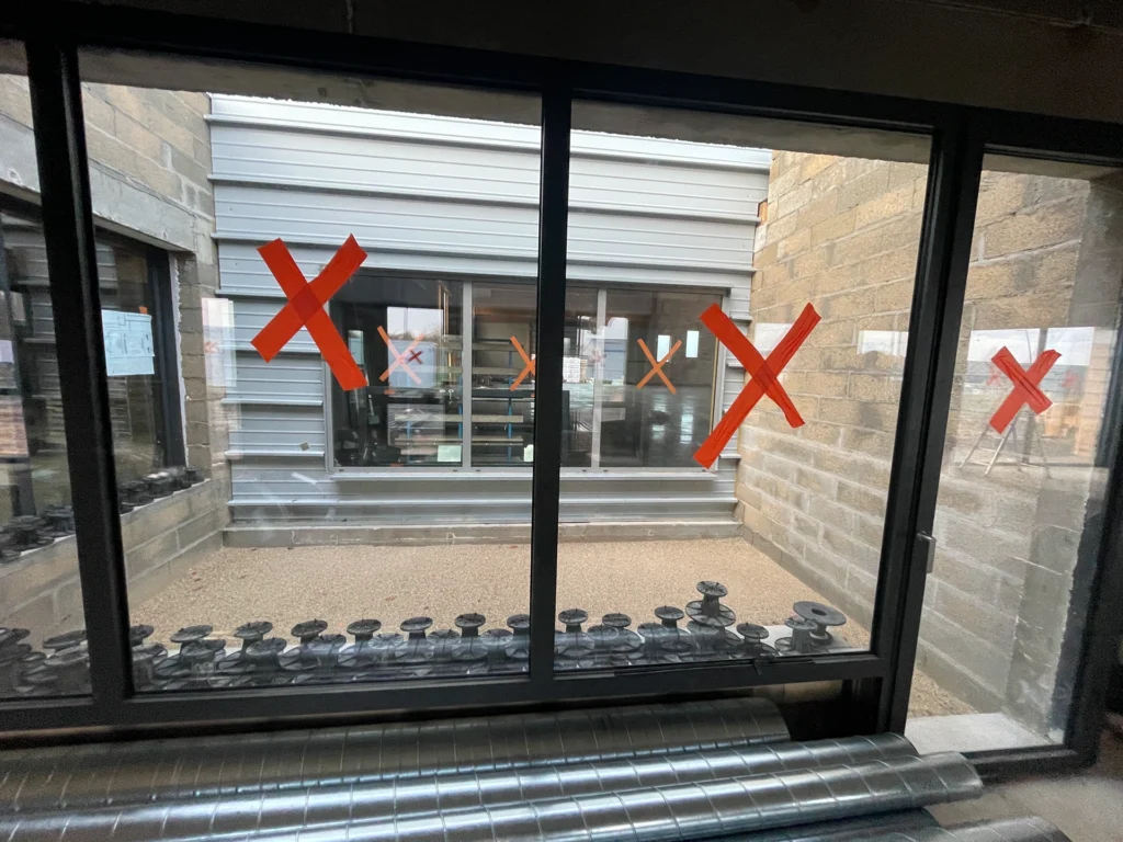De grandes marques X rouges faites avec du ruban adhésif sont placées sur plusieurs fenêtres en verre d'un Chantier Hadecoville, vues de l'intérieur d'un bâtiment aux murs en béton et à la cour recouverte de gravier, évoquant un style scandinave à la fois brut et professionnel.