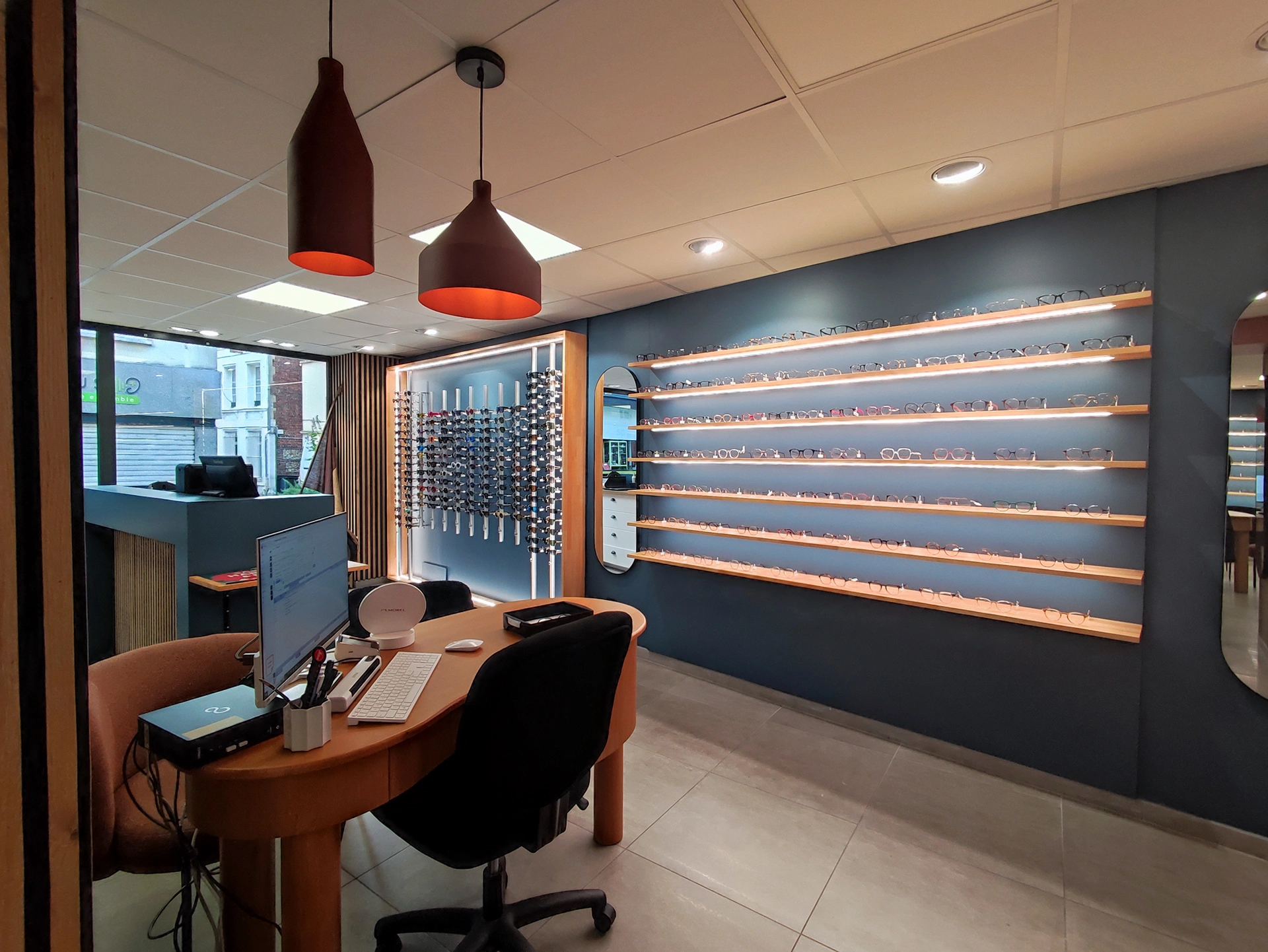 Une boutique professionnelle moderne de Hadecoville avec un bureau, un ordinateur et une chaise au premier plan. Des montures de lunettes sont soigneusement disposées sur des étagères le long d'un mur bleu, avec des lampes suspendues au-dessus et une fenêtre donnant sur le style contemporain de la rue.