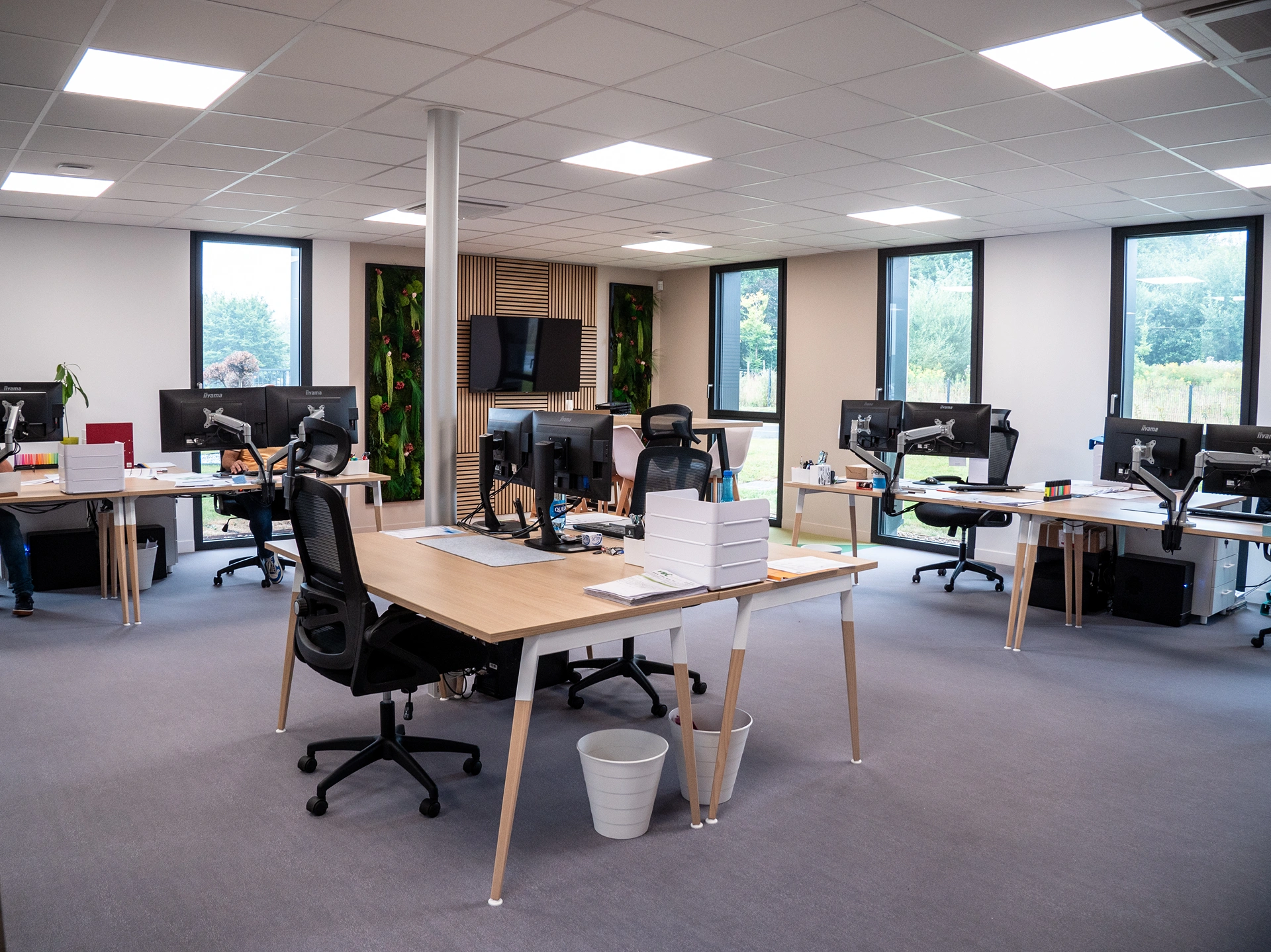 Bureau professionnel moderne à aire ouverte à Hadecoville, avec une touche de style scandinave : plusieurs bureaux, chaises, ordinateurs et écrans. De grandes fenêtres inondent de lumière cet espace de travail ordonné et calme, tandis que des plantes vertes apportent de la couleur et de la fraîcheur.