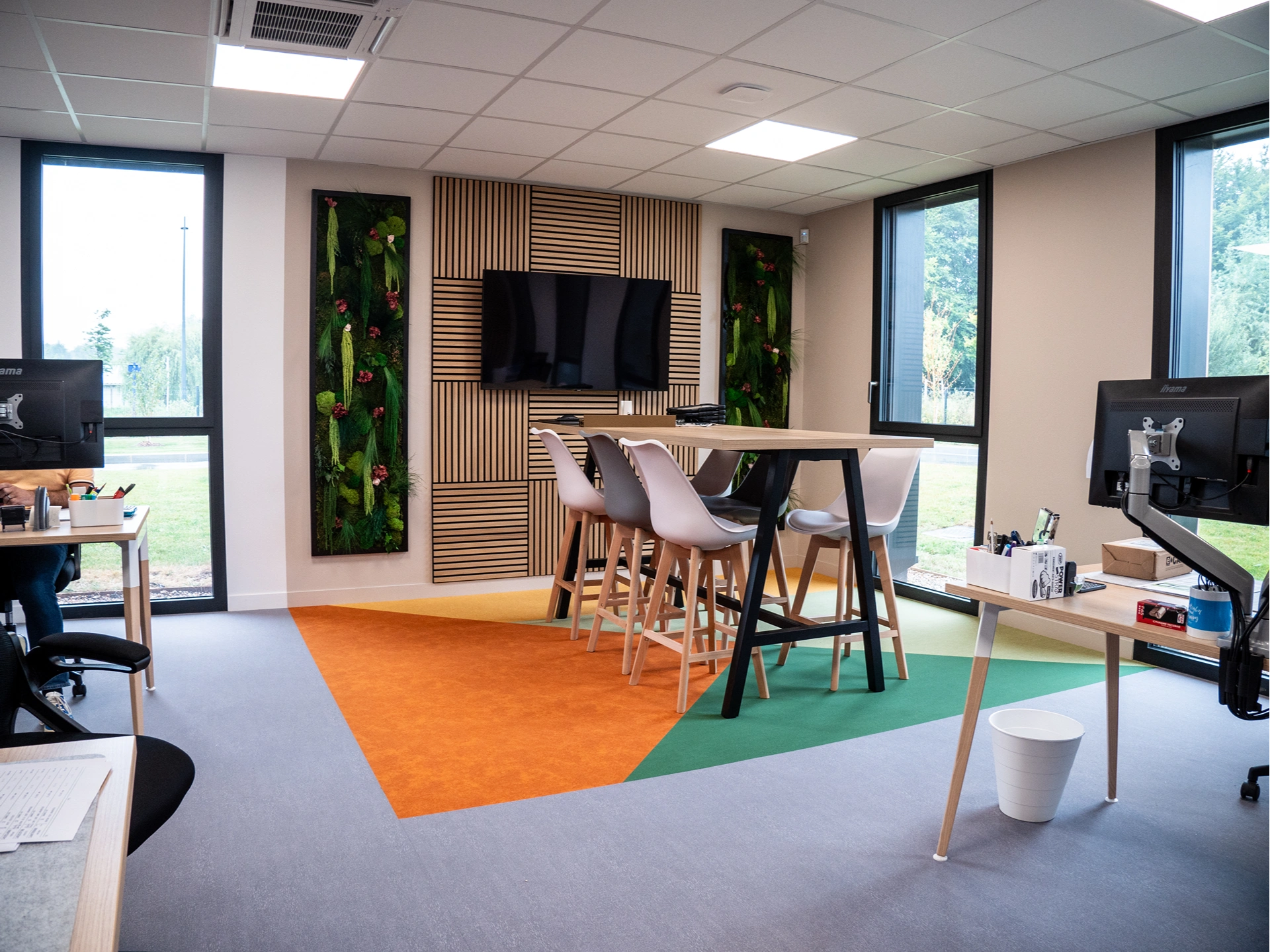 Bureau moderne Hadecoville professionnels avec une table haute et des chaises, une télévision murale, des bureaux avec des ordinateurs, de grandes fenêtres et des panneaux de plantes vertes. Le sol présente des motifs de tapis géométriques colorés dans le style scandinave.