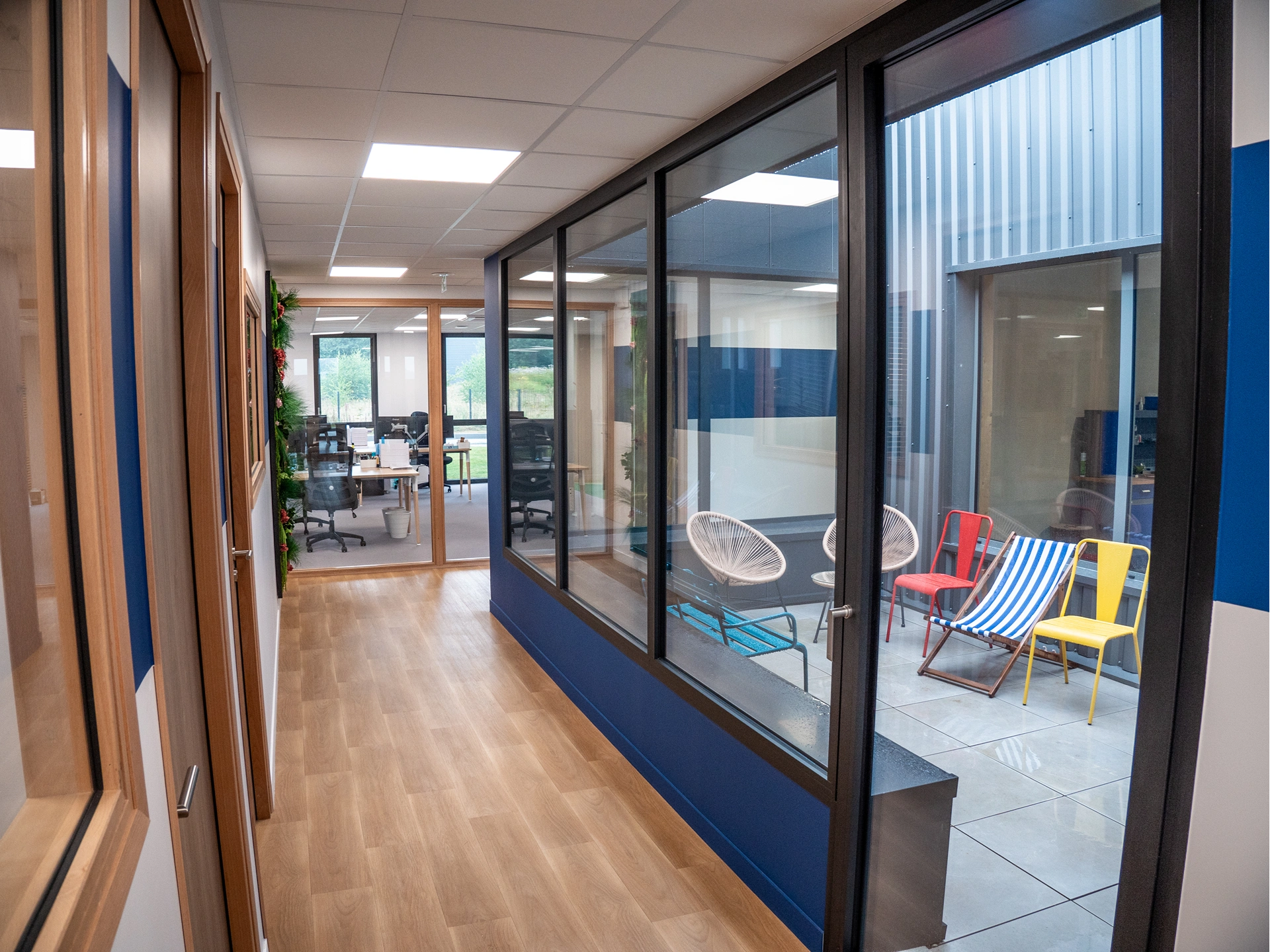 Couloir avec sol en bois et grandes fenêtres donnant sur une petite cour avec des chaises colorées. Au bout, les bureaux professionnels Hadecoville de style scandinave sont visibles à travers des parois vitrées, offrant un espace de bureau moderne.