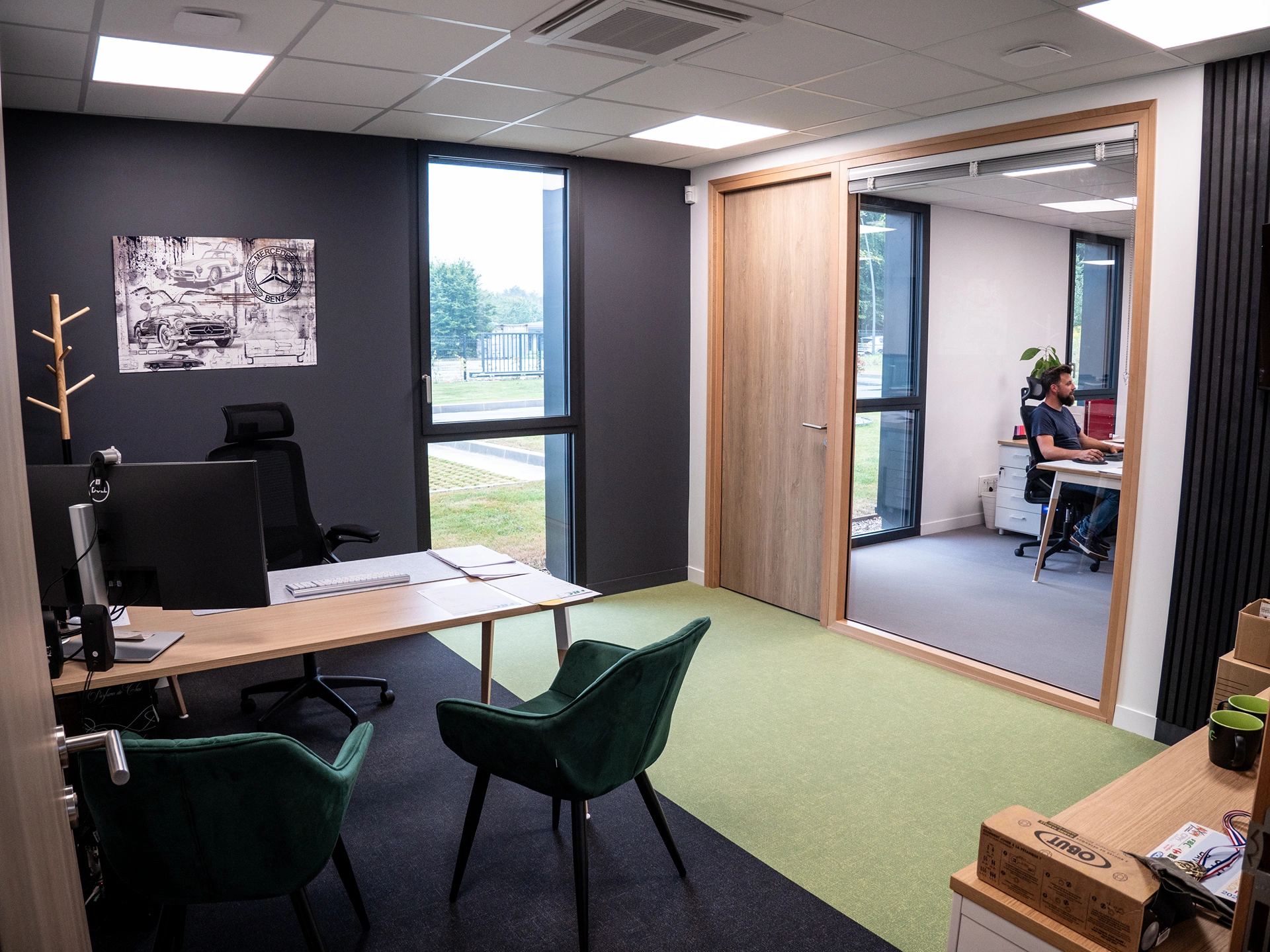 Un bureau moderne à Hadecoville bureaux professionnels style scandinave comprend un bureau, deux chaises vertes, un ordinateur et des œuvres d'art murales. À travers une paroi vitrée, quelqu'un travaille à proximité tandis que de grandes fenêtres remplissent l'espace de lumière naturelle.
