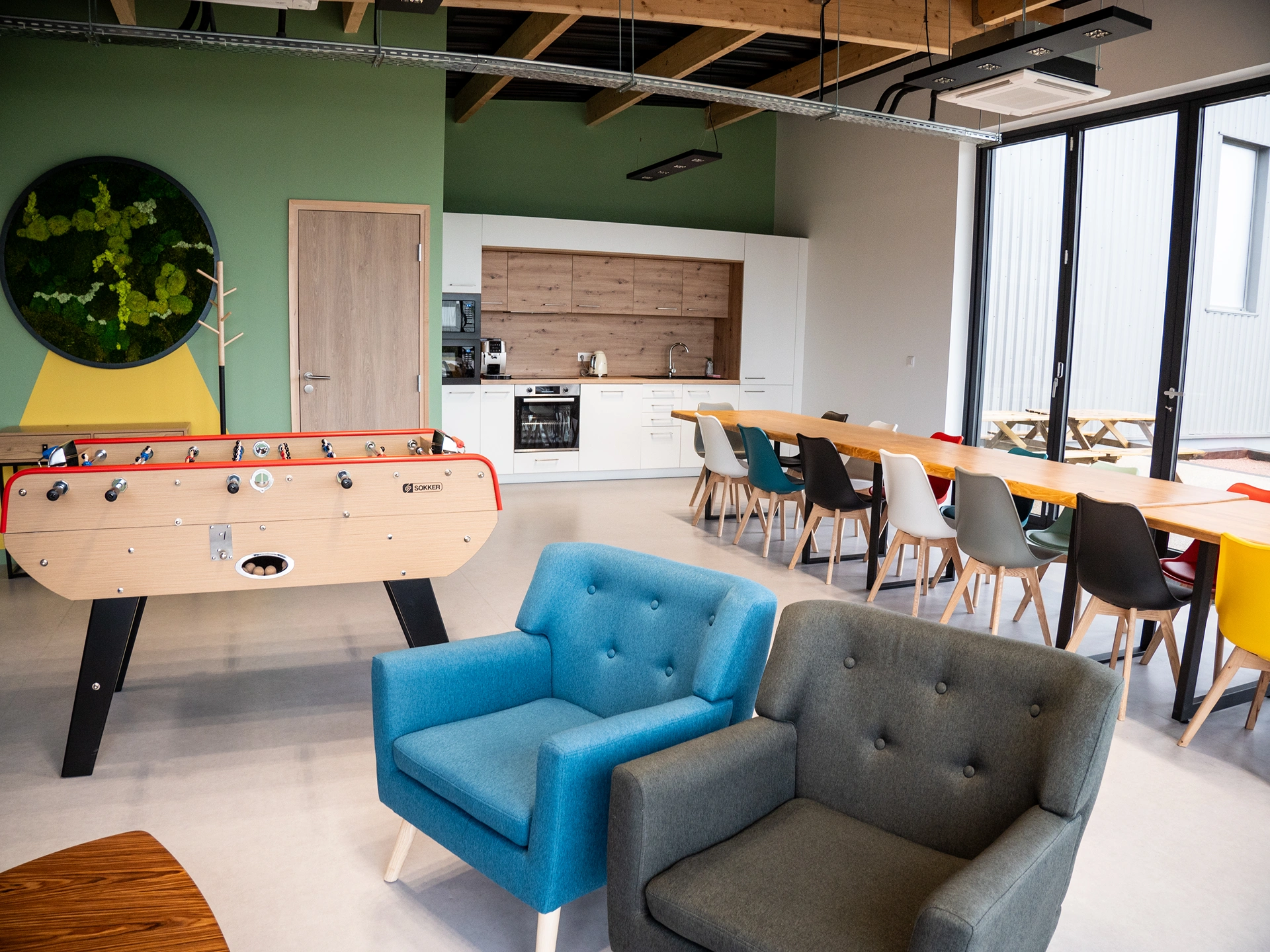 Salon de bureau moderne avec un baby-foot, des chaises de salle à manger colorées autour de tables en bois, un coin cuisine, deux fauteuils et de grandes fenêtres laissant entrer la lumière naturelle. Un mur vert avec des plantes décore l'espace.