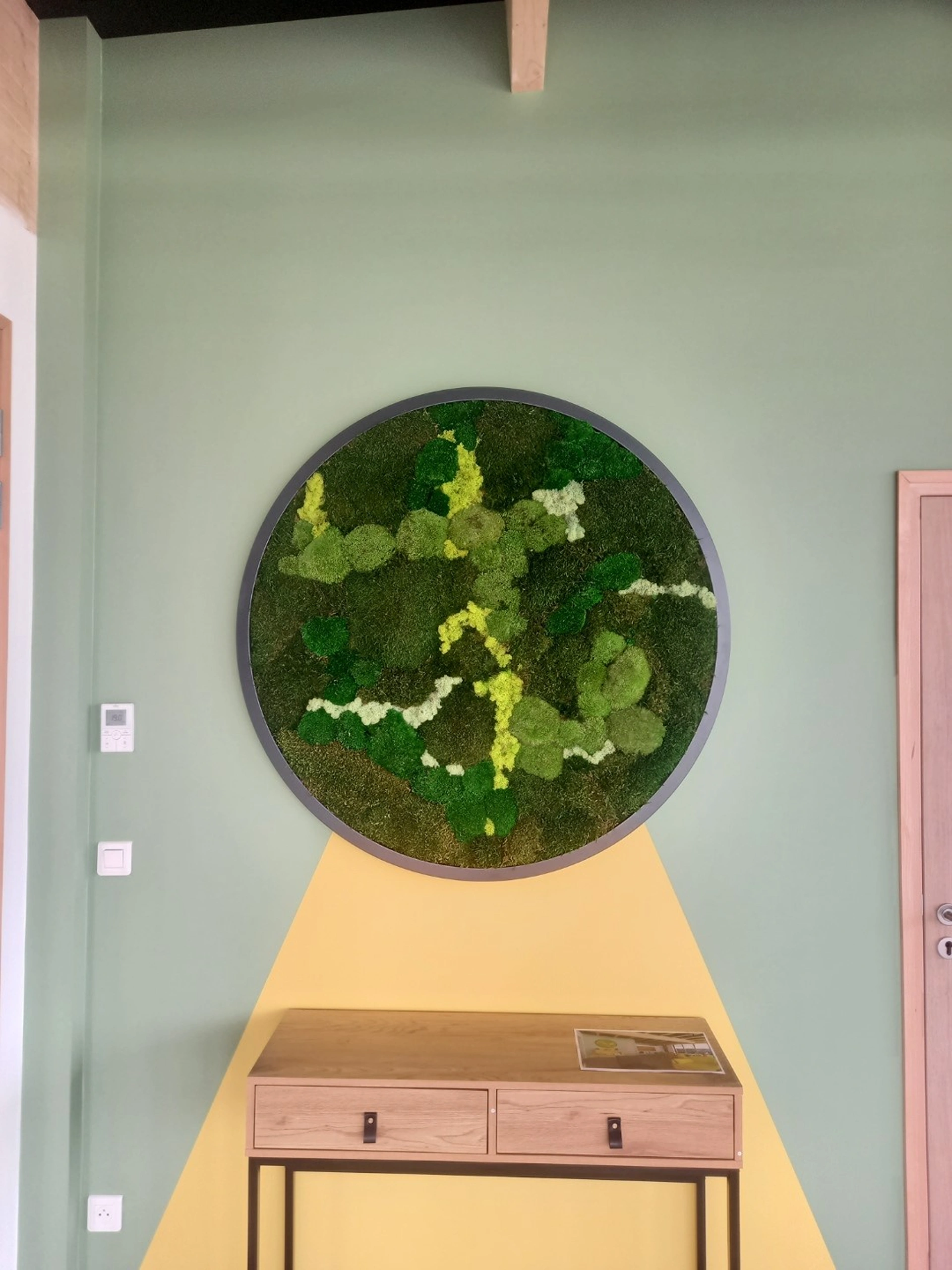 Une œuvre d'art murale ronde avec des mousses vertes et jaunes, inspirée des bureaux professionnels de style scandinave d'Hadecoville, est suspendue au-dessus d'une table console en bois. Le mur vert clair est orné d'un triangle jaune sous le cercle, évoquant la lumière qui brille. Un magazine est posé sur la table.