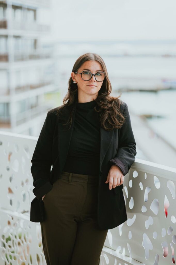 Une femme aux longs cheveux bruns, portant des lunettes, un blazer noir et un pantalon sombre, se tient sur un balcon aux balustrades décoratives. Elle regarde l'appareil photo avec assurance, avec des bâtiments et de l'eau en arrière-plan.